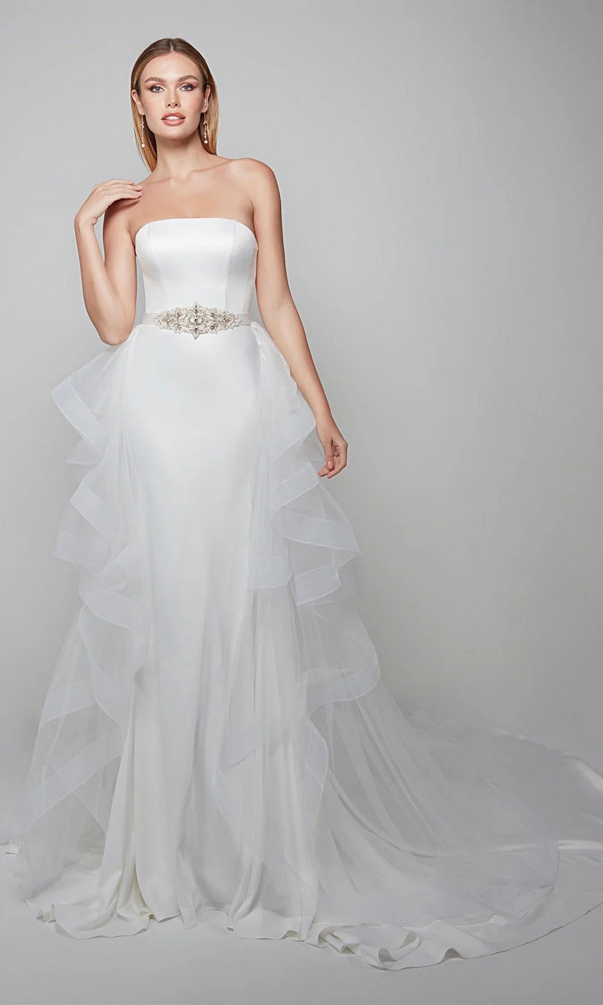 A woman wears the Alyce 7025 long white dress in diamond white, featuring a strapless satin bodice, jeweled belt, sheer ruffled sides, and a layered tulle overskirt—perfect for a wedding reception against a plain background.